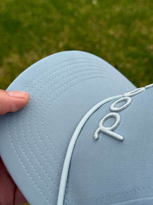 A close-up of a light golf cap held by a hand, showing white embroidered lettering on the front and a white piping detail along the brim, with a blurred green grass background.